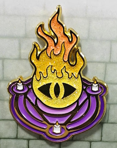 A glittering enamel pin featuring a stylized flame and an eye design in orange and purple colors, representing the Warlock character class.