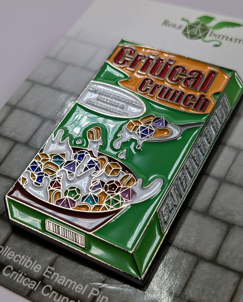 A colorful enamel pin depicting a box of Critical Crunch breakfast cereal with a spoon, now with berries and malted milk. Milk not included.