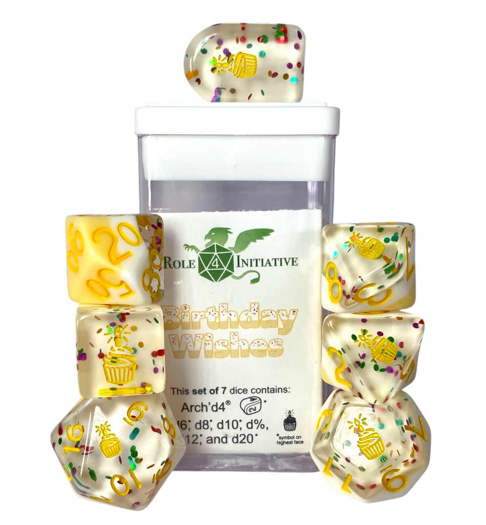 This photo is a set of 7 dice pictured with the box and label. It shows that the dice are layered with yellow on the bottom, white in the middle, and a clear layer with "confetti" glitter on top, and dark yellow numbers. The highest number on all the dice except the d% is replaced by a symbol; for this set, it's a cupcake with a candle.