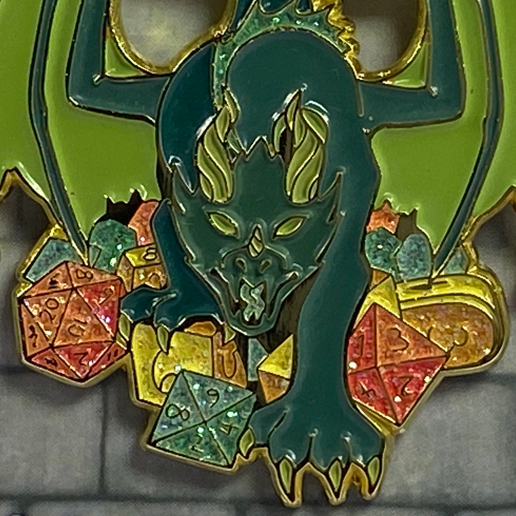 Enamel pin with a dragon design and polyhedral dice on a green background